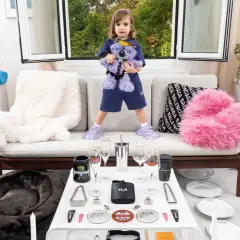 Dziewczynka z pluszowym misiem Balenciaga, otoczona zabawkami i akcesoriami tej marki. Czy to balenciaga co to za firma? Tak, to znany dom mody.