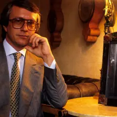 Mężczyzna w okularach, w garniturze, z krawatem, siedzi obok ramki ze zdjęciem palącego mężczyzny. To projektant Gucci.