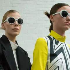 Dwie modelki w okularach w stylu miu. Jedna w czarnym płaszczu, druga w żółto-czarnej bluzce z geometrycznym wzorem.