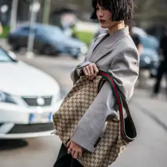 Kobieta w szarym żakiecie trzyma torbę z monogramem Gucci, jakby to był certyfikat autentyczności.