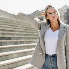 Chiara Ferragni w szarej marynarce w kratę i jeansach, z ręką we włosach, na tle schodów.