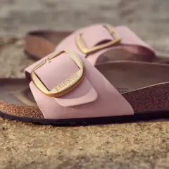 Różowe klapki Birkenstock z złotą klamrą, leżące na kamiennym podłożu.