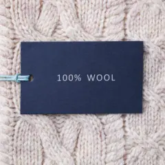 Ciemnoniebieska metka z napisem "100% WOOL" na jasnym, plecionym swetrze z grubej wełny jagnięcej.