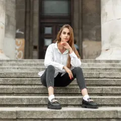 Eleganckie mokasyny to idealne buty do szarych spodni. Kobieta w białej koszuli i czarnych jeansach siedzi na schodach.