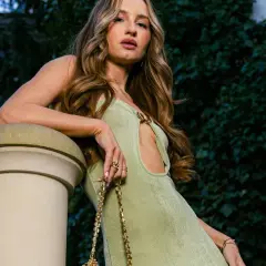 Młoda influencerka w zielonej sukience z wycięciem, oparta o kamienną balustradę, pozuje z torebką na złotym łańcuszku.
