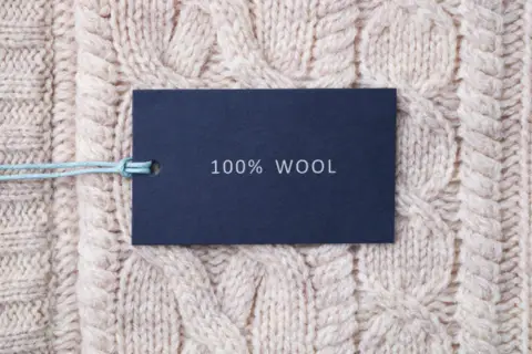 Ciemnoniebieska metka z napisem "100% WOOL" na jasnym, plecionym swetrze z grubej wełny jagnięcej.