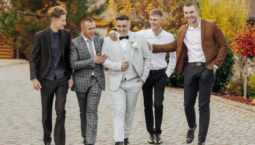 Pięciu mężczyzn w eleganckich strojach na wesele jako goście. Jeden w jasnej, dwurzędowej marynarce, inny w brązowej.