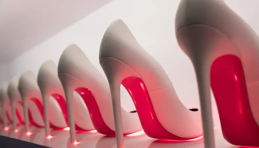 Rząd eleganckich, jasnych szpilek z czerwonymi podeszwami, przypominających kultowe Louboutiny, lśni w blasku neonowego światła.