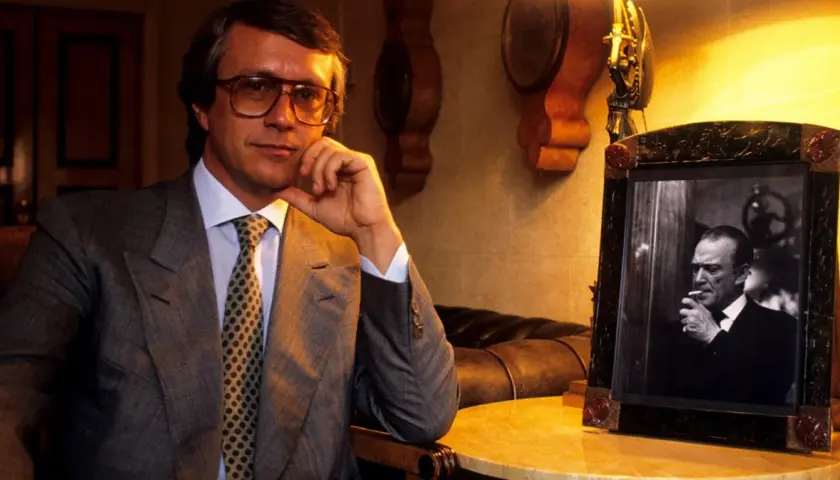 Mężczyzna w okularach, w garniturze, z krawatem, siedzi obok ramki ze zdjęciem palącego mężczyzny. To projektant Gucci.