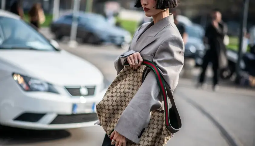 Kobieta w szarym żakiecie trzyma torbę z monogramem Gucci, jakby to był certyfikat autentyczności.