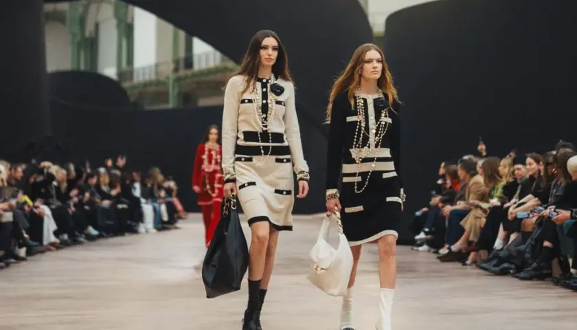 Dwie modelki prezentują kreacje francuskiego domu mody na wybiegu. Eleganckie sukienki w stylu Chanel, bogata biżuteria i torebki.