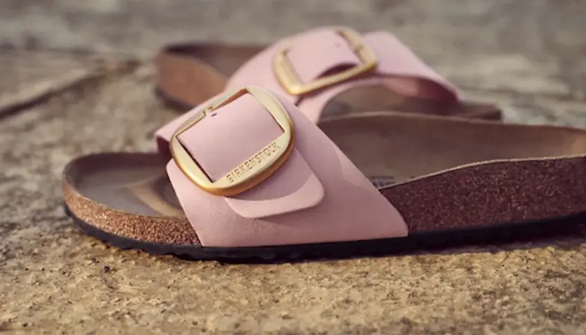 Różowe klapki Birkenstock z złotą klamrą, leżące na kamiennym podłożu.