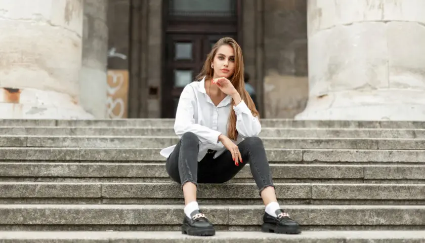 Eleganckie mokasyny to idealne buty do szarych spodni. Kobieta w białej koszuli i czarnych jeansach siedzi na schodach.