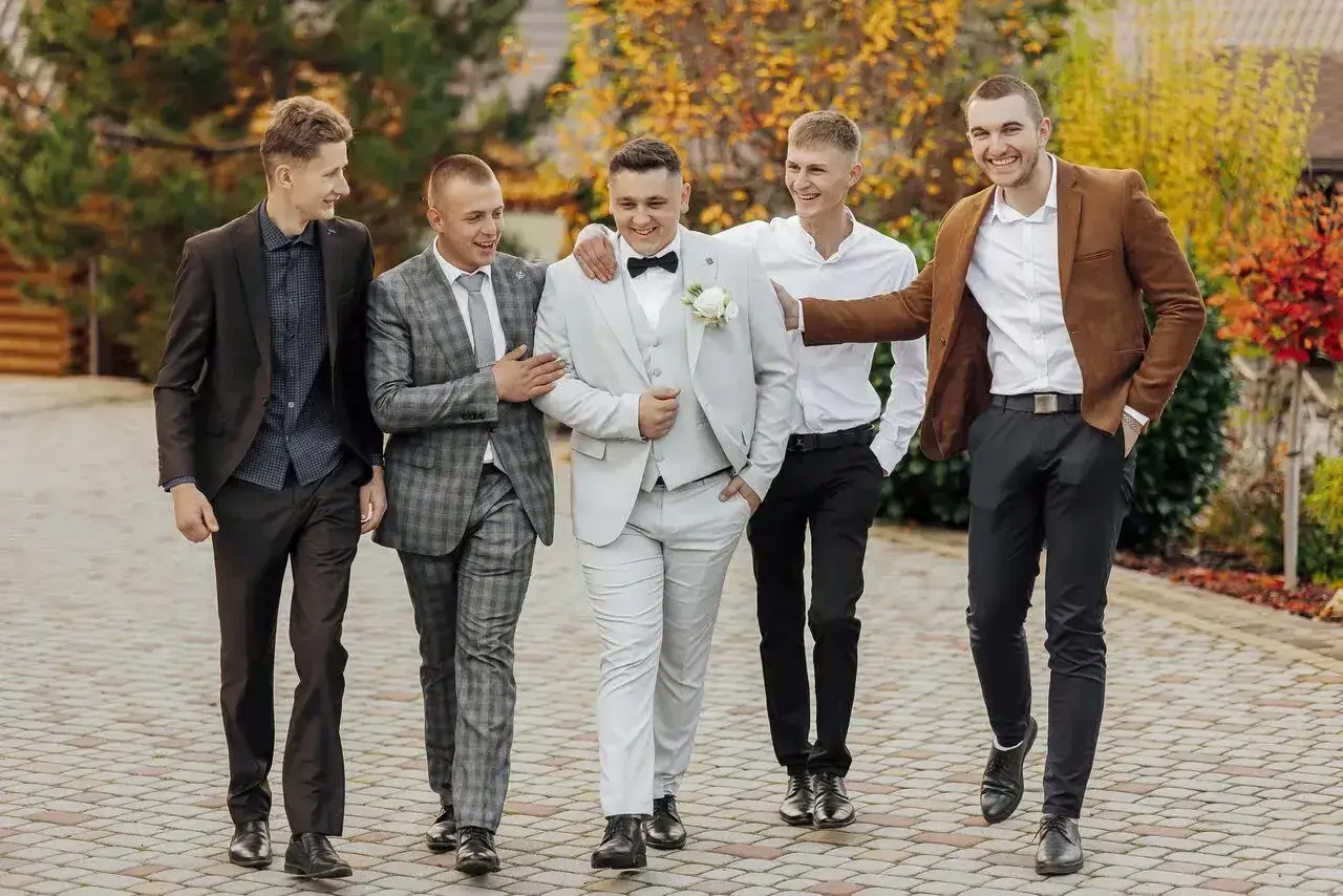 Pięciu mężczyzn w eleganckich strojach na wesele jako goście. Jeden w jasnej, dwurzędowej marynarce, inny w brązowej.