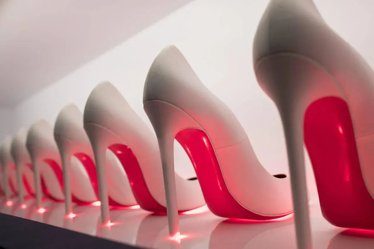 Rząd eleganckich, jasnych szpilek z czerwonymi podeszwami, przypominających kultowe Louboutiny, lśni w blasku neonowego światła.