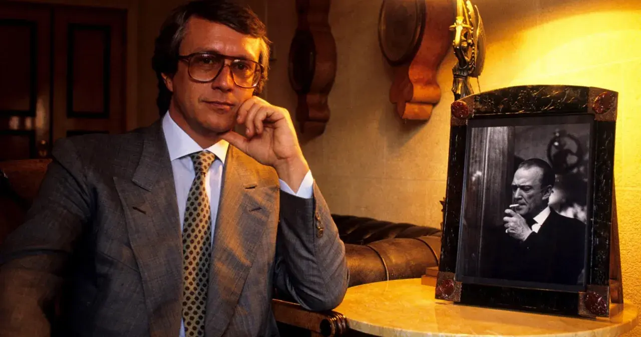 Mężczyzna w okularach, w garniturze, z krawatem, siedzi obok ramki ze zdjęciem palącego mężczyzny. To projektant Gucci.