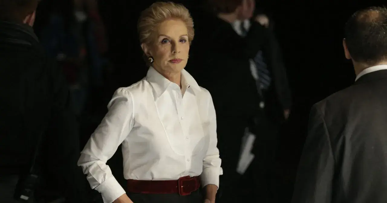 Elegancka Carolina Herrera w białej koszuli i czerwonym pasie, otoczona przez ludzi w ciemnych strojach.
