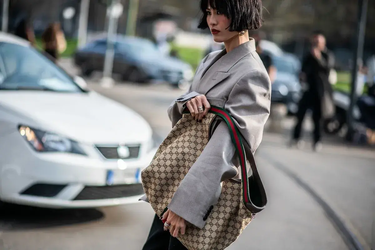 Kobieta w szarym żakiecie trzyma torbę z monogramem Gucci, jakby to był certyfikat autentyczności.