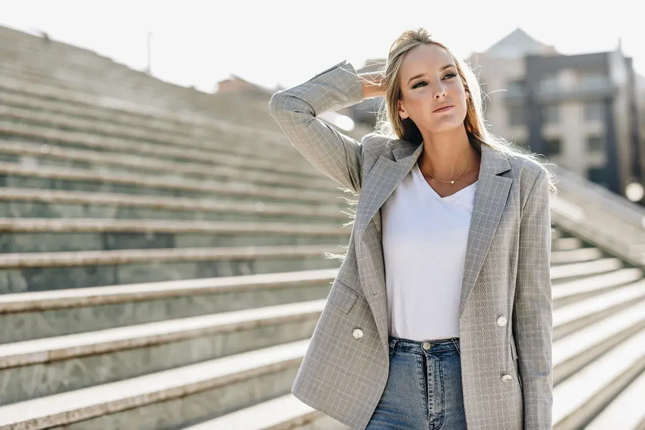 Chiara Ferragni w szarej marynarce w kratę i jeansach, z ręką we włosach, na tle schodów.