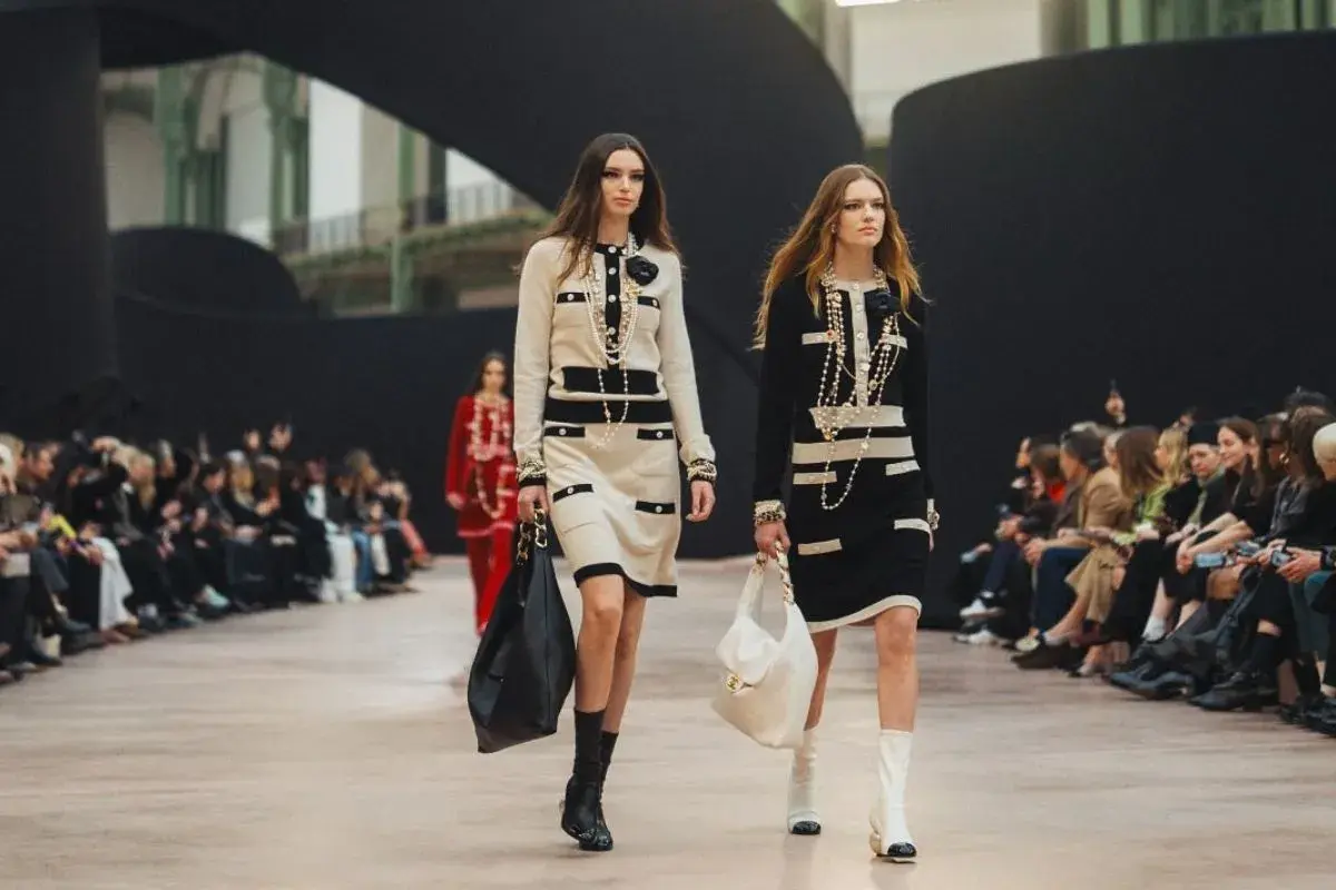 Dwie modelki prezentują kreacje francuskiego domu mody na wybiegu. Eleganckie sukienki w stylu Chanel, bogata biżuteria i torebki.