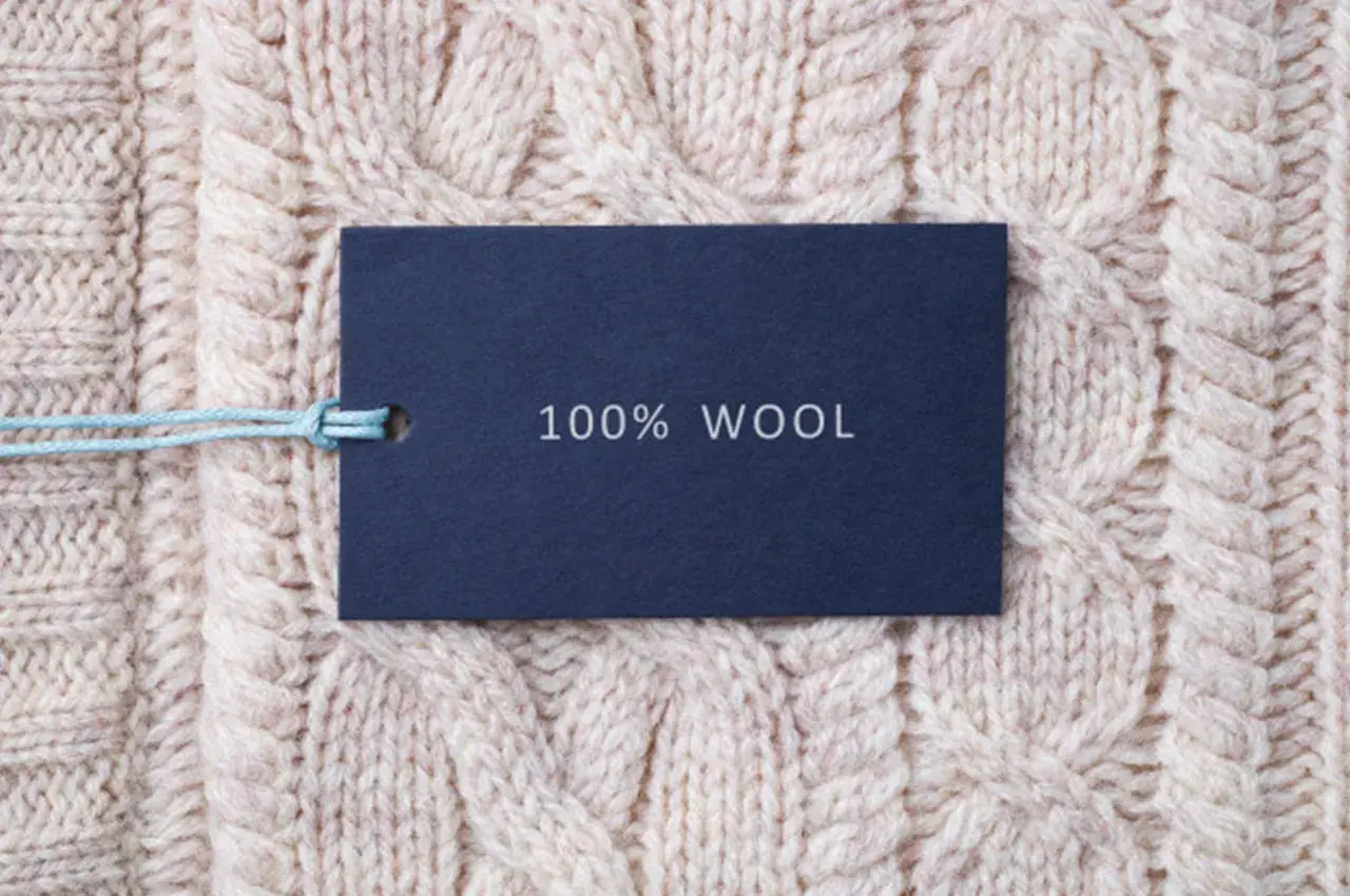 Ciemnoniebieska metka z napisem "100% WOOL" na jasnym, plecionym swetrze z grubej wełny jagnięcej.