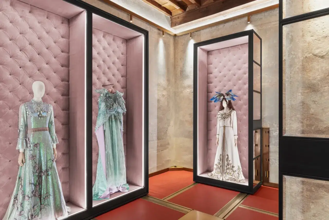Wystawa kreacji projektanta Gucci: błyszczące suknie i ekstrawaganckie nakrycia głowy w różowych gablotach.