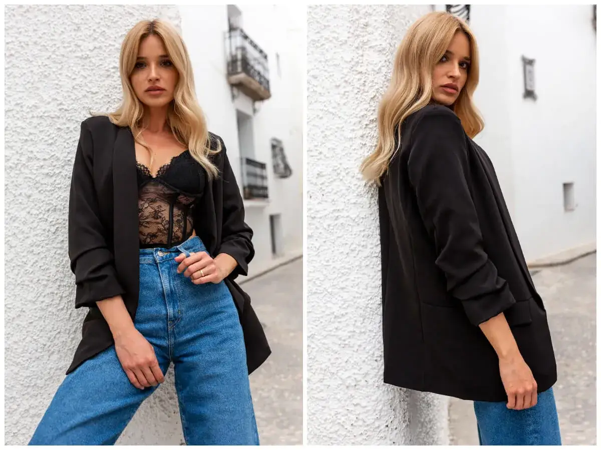 Eleganckie stylizacje z jeansami: czarny blazer, koronkowy top i jeansy. Kobieta pozuje na tle białej ściany.