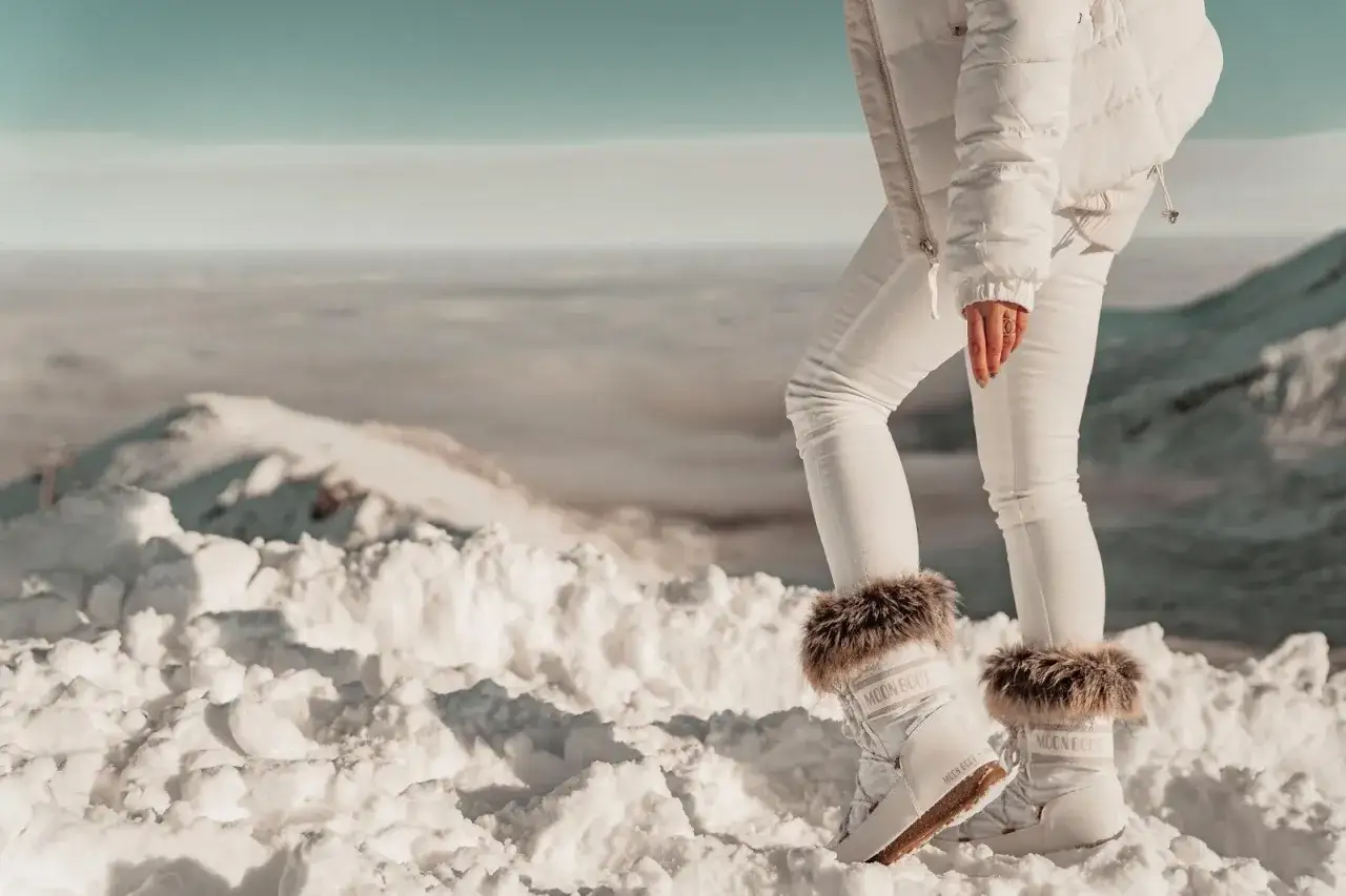 Białe buty Moon Boot z futerkiem na śniegu, idealne na après-ski. Widok na góry i chmury.
