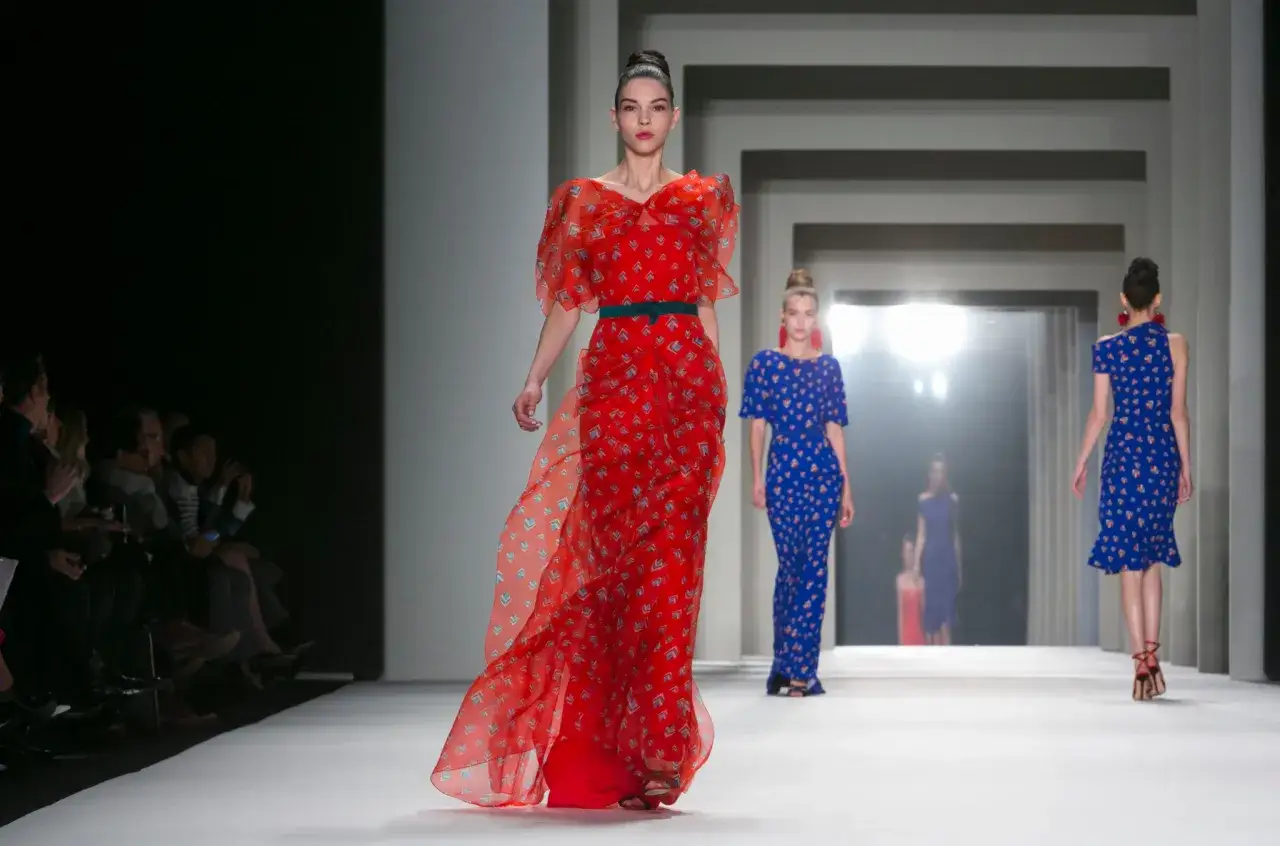 Modelka w czerwonej sukni z wzorem w romby, prezentująca kreację od Herrera, kroczy po wybiegu.